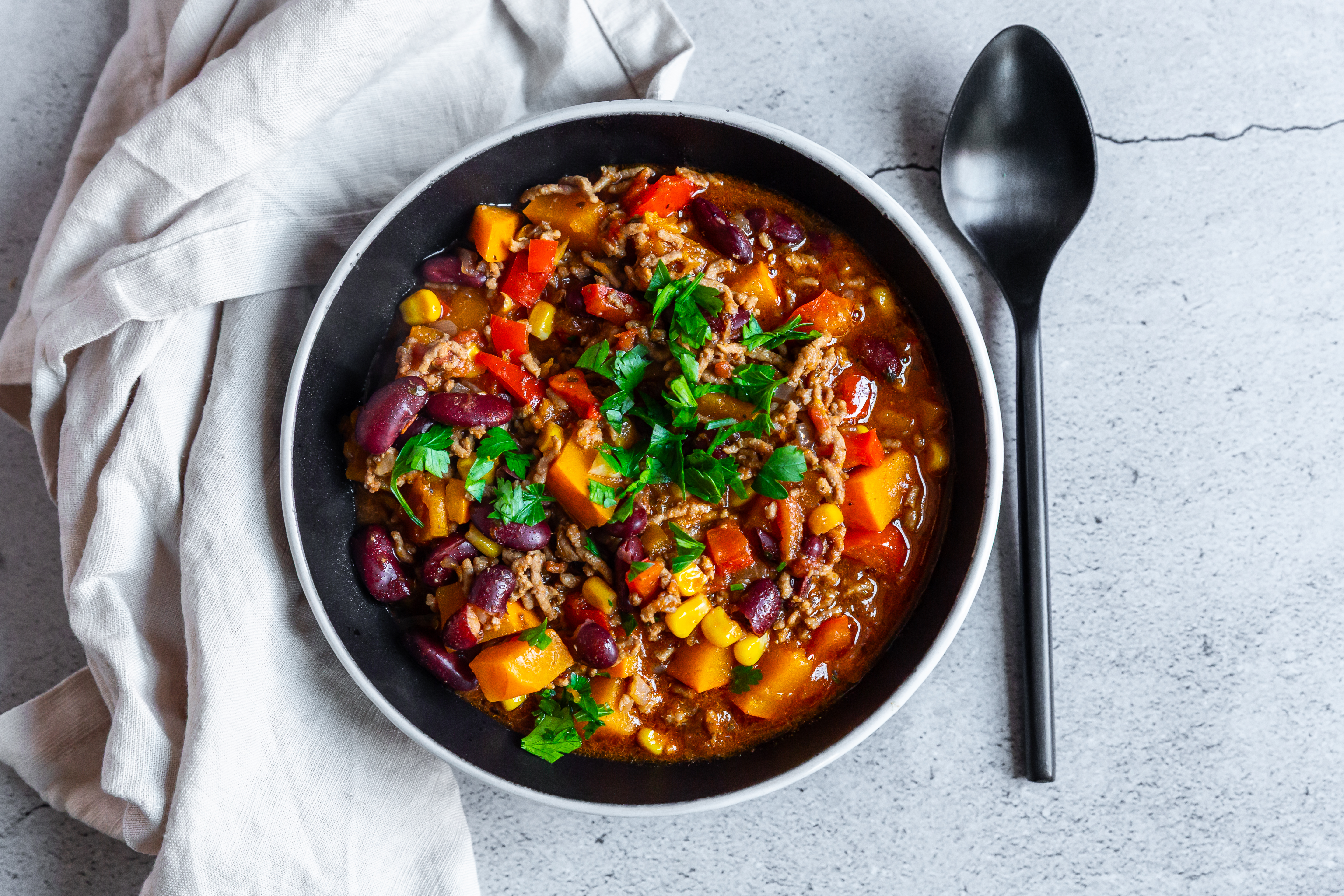 Chili con Carne mit Hokkaidokürbis, Kidneybohnen, Mais und roter Paprika in einer dunklen Schüssel, garniert mit frischer Petersilie.